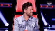 a man in a denim jacket is sitting in a chair on a stage and smiling .