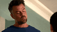 a man with a beard is wearing a blue shirt and talking to another man in a room .