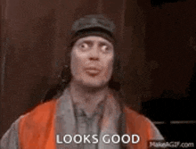 a man wearing a hat and an orange vest is making a funny face and says `` looks good '' .