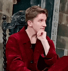 a man in a red coat is sitting in a chair with his hands on his chin .