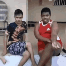 two young men are sitting next to each other on the floor . one of the men is wearing a red adidas shirt .