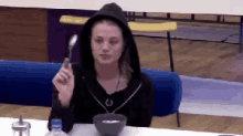 a woman in a black hoodie is sitting at a table with a bowl of food and a spoon in her hand