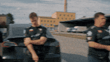 two men standing in front of a car with one wearing a t-shirt that says ' rs ' on it