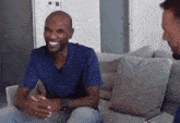 a man in a blue shirt is smiling while sitting on a couch talking to another man