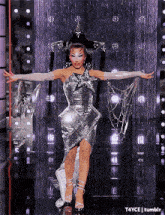 a woman in a silver dress and white gloves is standing on a stage with her arms outstretched .