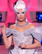 a drag queen wearing a silver dress and a necklace is standing in front of a pink wall
