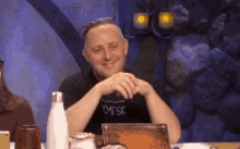 a man is smiling while sitting at a table with a laptop and a water bottle .