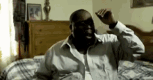 a man in a white shirt is sitting on a bed with his hands in the air .
