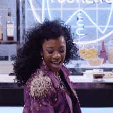 a woman in a purple jacket is smiling in front of a neon sign that says ' u '