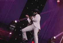 a man in a white suit and hat is dancing on a stage with purple lights behind him