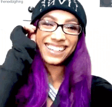 a woman with purple hair and glasses is smiling while wearing a black hat .
