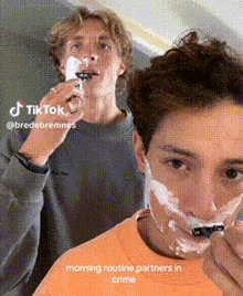 two young men are shaving their faces in a tiktok video . one of the men is wearing an orange shirt .