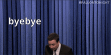 a man in a suit and tie says byebye in front of a blue curtain