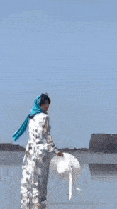 a woman in a white hat and blue scarf is walking on the beach