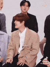 a group of young men are sitting in a room and one of them is wearing a brown jacket .