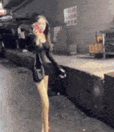 a woman in a black dress is walking down a street while holding a cell phone .