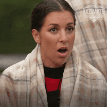 a woman with a plaid scarf around her neck looks surprised with a canada 's ultimate challenge logo behind her