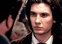 a man with long hair and a white shirt looks at another man