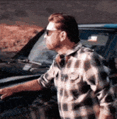 a man wearing sunglasses and a plaid shirt stands next to a car
