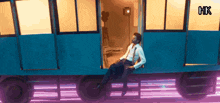 a man sits on the side of a blue train with hk written on the bottom right