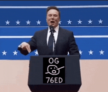 a man in a suit and tie is giving a speech at a podium that says og 76ed on it