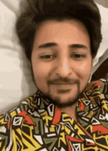 a man with a beard is wearing a colorful shirt and smiling while laying on a bed .