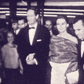 a man in a tuxedo and a woman in a white dress are walking in a crowd