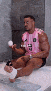 a man in a pink adidas shirt is sitting in a bathtub holding a ball