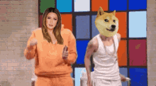a woman in an orange sweatshirt stands next to a man in a white tank top with a doge on his head