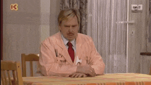 a man in a pink suit sits at a table with a tennis ball on the wall above him that says op12