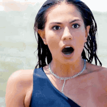 a woman wearing a blue one shoulder swimsuit and a silver chain necklace looks surprised