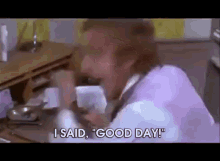 a man in a purple shirt is sitting at a table talking on a cell phone and saying i said good day .