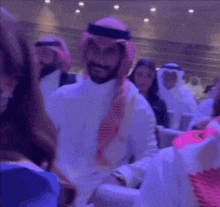 a man wearing a keffiyeh and a hat is sitting in a crowd of people and smiling .