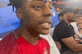 a man wearing a red shirt with a green collar sits in a stadium watching a soccer game