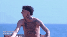 a shirtless man with a lot of tattoos on his chest is sitting on a ledge overlooking the ocean .