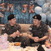 two men in military uniforms are sitting at a table with cakes and balloons in the background