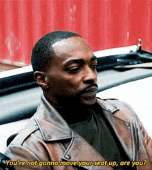 a man in a brown leather jacket is sitting in a car and talking to another man .
