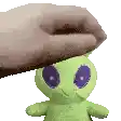 a green stuffed animal with purple eyes is being held by a hand .