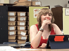 a woman sits at a desk with a tablet in front of a shelf with boxes labeled paper and daniela