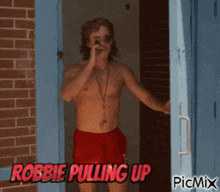 a shirtless man in red shorts is standing in front of a brick wall with the words robbie pulling up on the bottom