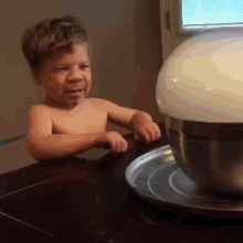 a shirtless little boy is sitting at a table looking at a white bowl