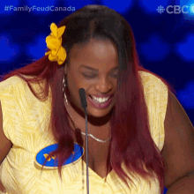 a woman with red hair and a yellow flower in her hair is smiling in front of a microphone with the words family feud canada behind her