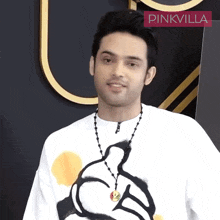 a man wearing a white shirt with a mickey mouse necklace is standing in front of a pinkvilla banner