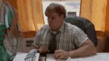 a man in a plaid shirt and tie is sitting at a desk in front of a green fan .