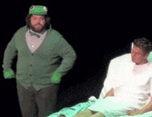 a man in a frog costume stands next to a man in a hospital bed .