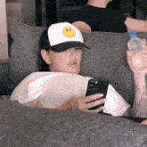 a woman wearing a hat with a smiley face on it is sitting on a couch looking at her phone