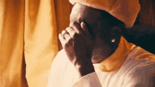 a man wearing a white shirt and a yellow hat is covering his face with his hands .