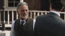 a man with gray hair and a beard talks to another man in a suit and tie