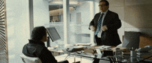 a man in a suit and tie stands next to another man in a chair