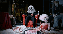 a man wearing an owl mask sits in a red chair next to a woman in a red dress